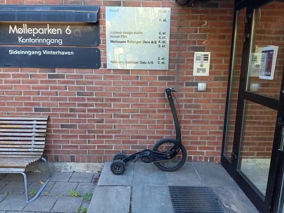 A Halkbike (a compact, foldable, standing bike with one front wheel and two rear wheels) in front of a brick wall, below a sign showing the location of offices of various businesses.