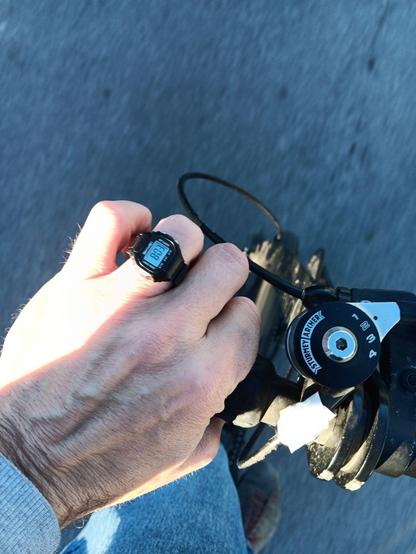 A view looking down at a hand holding very short handlebars. On the hand is a digital ring watch showing the time as 8:03.