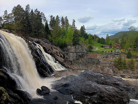 A waterfall. In the background are greenfields, farm buildings and some parked cars. 