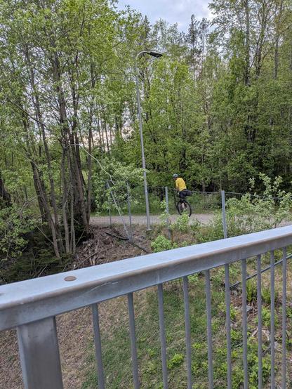 Man with a yellow shirt on a unicycle. The image is taken from a distance with tress all around.