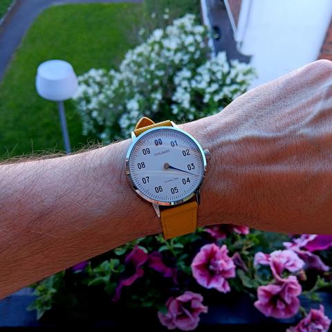 Decimal watch showing the time as 3.47 (~08:20AM). The watch has a white face with a single blue hand and a bright yellow strap. There are flowers in the background. 