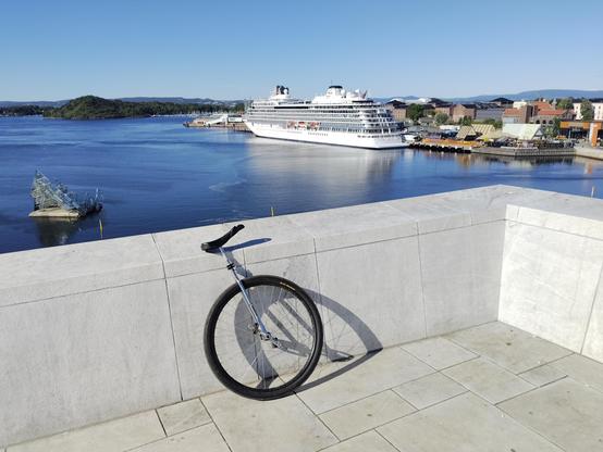 A sunny view of the Oslo fjord taken from the top of the Oslo Opera house. In the foreground is a white, smooth stone wall with a large wheeled unicycle leaning against it. In the background is the fjord with the glass sculpture, "She Lies" on the left and a lare cruise ship on the right.

On and there is no bin in the picture because Matt clearly stole it.