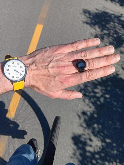 Looking down at my arm and wrist. I'm wearing a decimal watch and a digital ring watch. Below that it is possible to see the wheel of my unicycle and it is obvious I'm riding it while the picture is taken. 