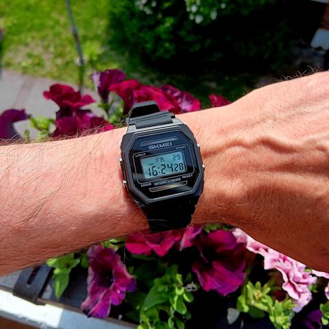 A simple, black digital watch on wrist. Flowers in the background. 