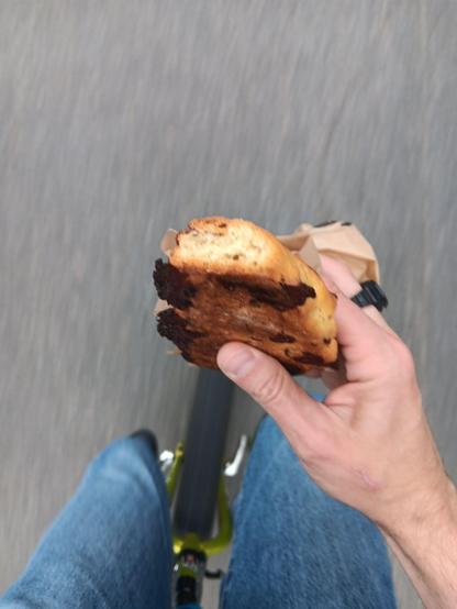 Unicyclist rider point of view looking down at their hand holding a chocolate scone. They are also wearing a ring watch and holding a paper bag. Below that you can see their legs and the frame and wheel of the unicycle.