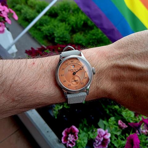 A watch with a salmon coloured dial and a gray strap. The dial only has two hands, minutes around the face and seconds in a sub dial. The hour is shown in a window, similar to a date window. In the background of the image are flowers and the edge of a pride flag.