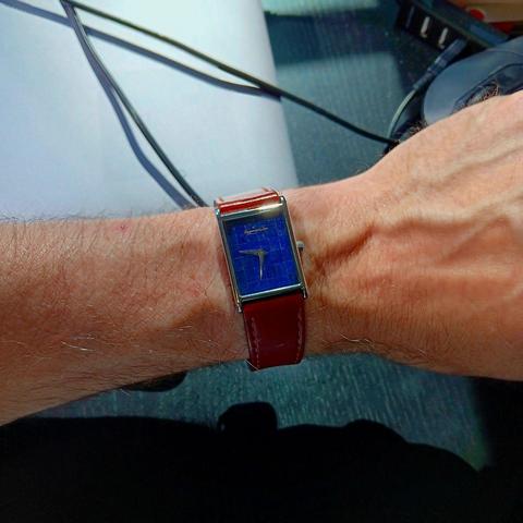 Rectangular watch, on writs with an intense blue dial, and no hour/minute markings. It has a brown leather strap. In the background is a black deskt with a mac laptop and a black mouse
