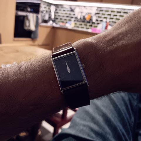 Rectangular watch showing the time as 12AM. Dark in the foreground on wrist, with the room lit up behind. 