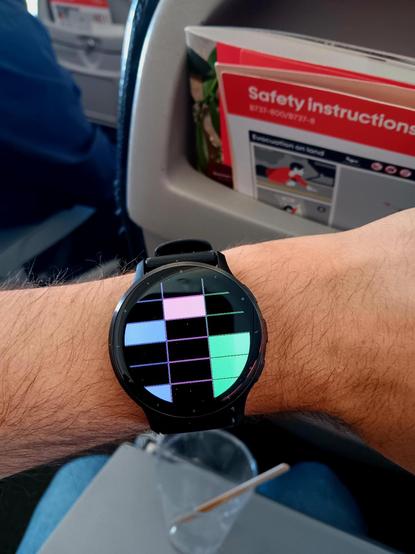 A watch on wrist, with a face encoding the time in binary (09:16 on the 7th day of the month). In the background you can see the back of a plane seat and a flight safety card. 