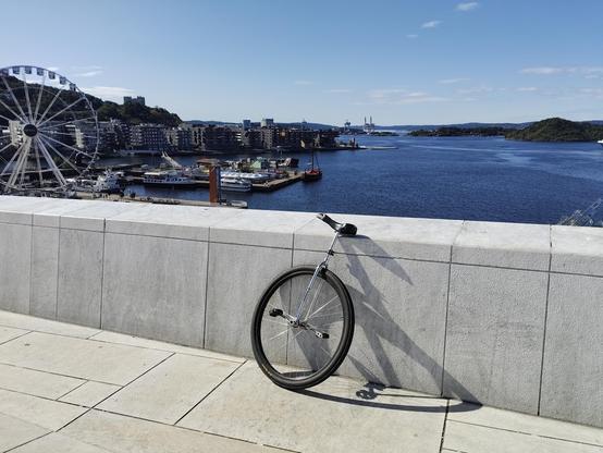 Picture from the top of the Oslo Opera house looking out at the fjord. A Ferris wheel is also visible to the right.

In the foreground is a large wheeled unicycle leaning on a white stone wall. 