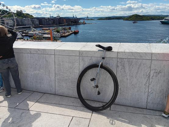 A view out over Oslo fjord from the top of Oslo Opera house. In the foreground is a large wheeled unicycle, leaning on a smooth, white, stone wall.
In the background is the harbour and a Viking style ship moored up.