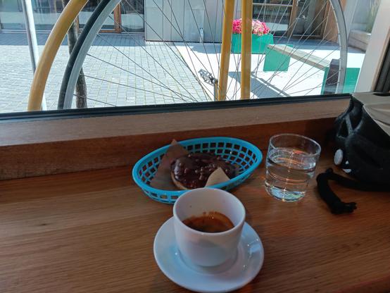 Don't and coffee resting on a table alongside water and a cycle helmet. There is a window behind, through which part of the frame and large wheel of a penny farthing bicycle can be seen.