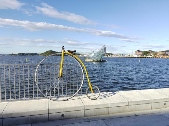 Yellow penny farthing bicycle, leaning on a metal fence beside the waterfront, with the glass sculpture She Lies floating in the Oslo Fjord in the background.