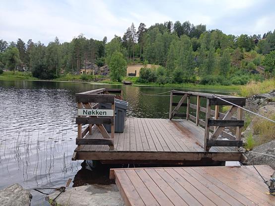 Small, wooden, cable ferry, viewed from to dock, looking out over the river. The ferry is labelled, Nøkken. Trees and the other dock on the other side. 