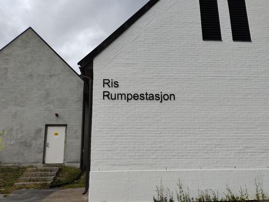 Building with a brick wall painted white, with white lettering that appears to read, "Ris Rumpestasjon". 