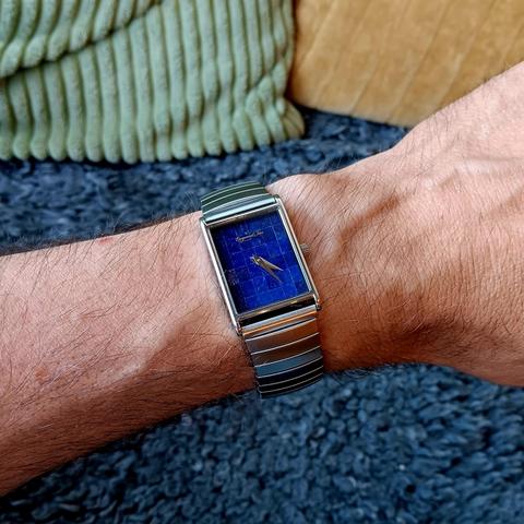 A 'tank' style rectangular watch with a deep blue dial made of smaller rectangles of lapis lazuli. Worn on a brushed steel bracelet, on wrist. In the background is a grey, woollen sheep skin, along with the edges of a green cushion and a yellow cushion. 