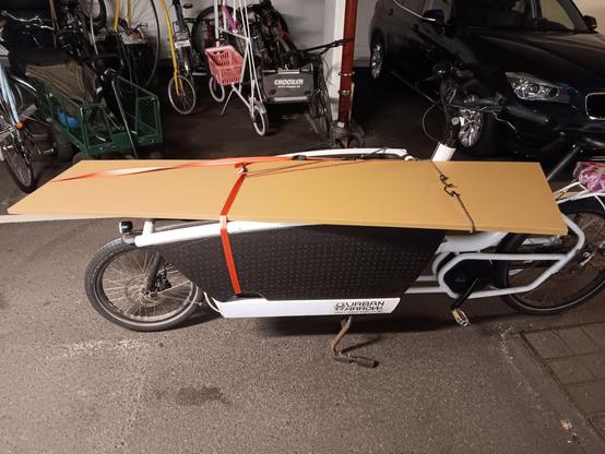 A cargo bike with a long cardboard box strapped on top. In the background are more bikes and a parked car.