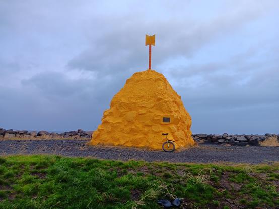 A large yellow mound of rock and concrete, with a yellow metal "flag" on top (it Suðurnesvarða, "the cairn/beacon of Suðurnes" and surved as a navigational waymarker). Leaning against it is a unicycle on black dirt, with grass in the foreground. The sky is gray and cloudy with small bits of light blue showing through. 
