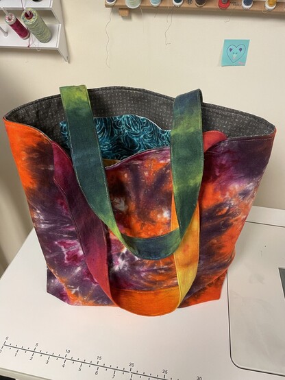 View of bag from purple/orange swirl side with green strap draped across and purple/orange strap hanging down. You can see the grey lining fabric and a blue wave pocket.