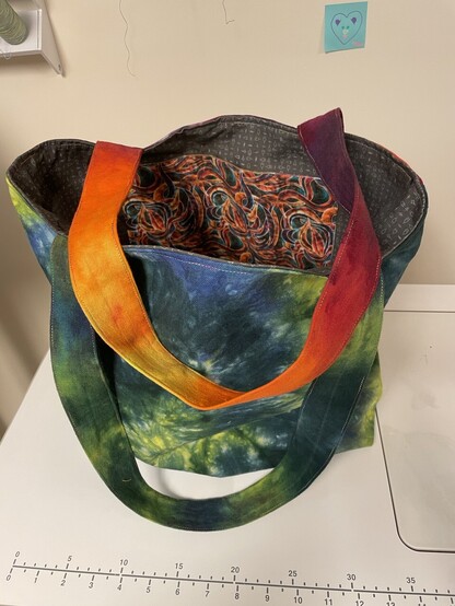 Green swirl side with orange/purple strap draped across the top and green strap hanging down in front. You can see a pocket made of rainbow wave fabric and the grey interior fabric.