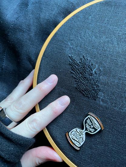 Lia’s fingers show an approximately 1.5 x 3 inch section of black on black whipped backstitch that looks a bit like flames, but if you’ve been following this piece you’ll know it’s pubic hair. It’s definitely pubic hair. An hourglass-shaped needle minder that reads “this is taking forever” is attached to the cloth. 