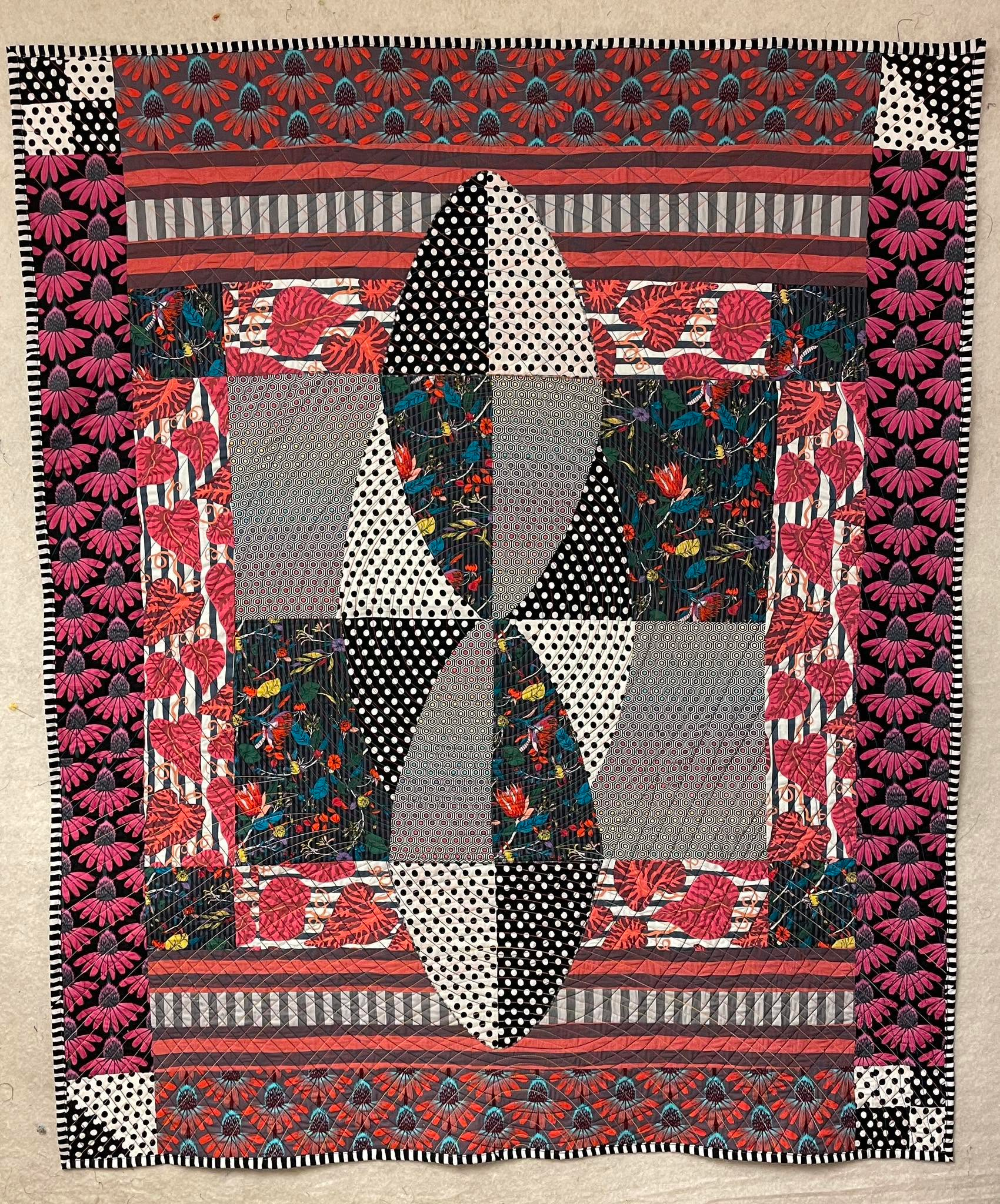 A 4ft by 5ft quilt. In the center are two ovals made from the same four fabrics, but flipped positionally. There are black polka dots on white and white on black on the outer portions and a black floral and a grey ombre hexagonal rainbow print on the inside. It is on top of a square that has a central oval made from the polka dots. The rest of that square is filled in with the grey hexagonal rainbow print and the black floral. Then there is a border of with large pink and orange leaves on a blue and white stripe and the black floral in the corners. At the top and bottom of this square is a sort of "railroad" motif with orange and grey stripes running horizontally and dark grey and light grey strips running vertically. On the top and bottom is an orange and blue print of cone flowers and on the sides is a black and pink version of the same print. At the top left and bottom right are little squares made of four patches of the polka dots and the top right and bottom left are two triangles of the polka dots. It is bound with a black and white striped fabric, but the stripes are a little wonky.