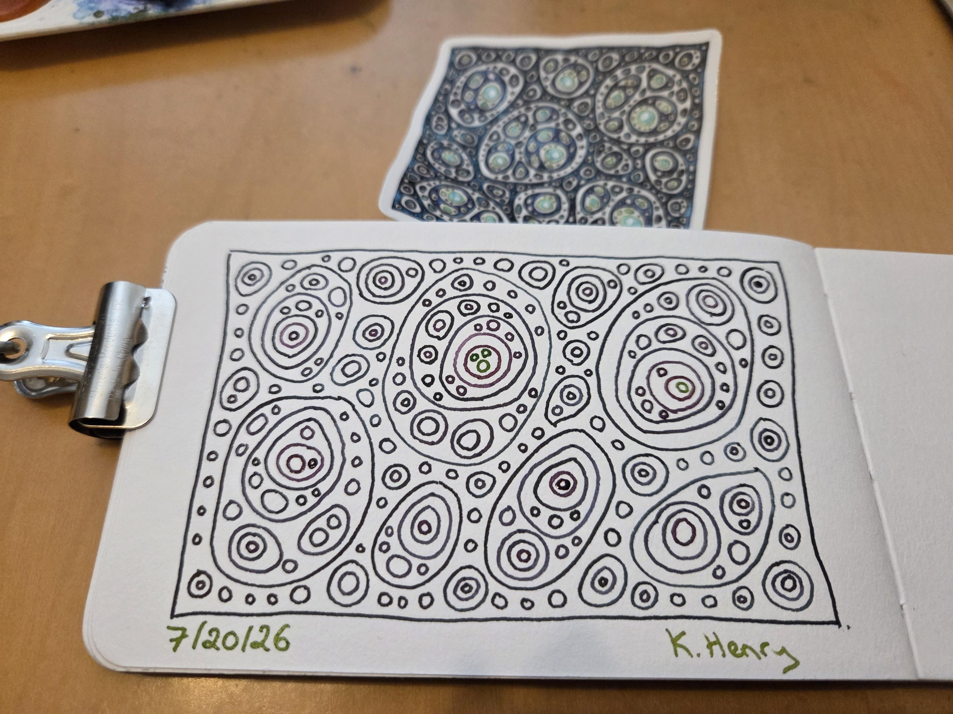 Hand drawn generative/procedural art in ink on an open page of my sketchbook. The abstract pattern looks a bit like something under a microscope in Biology lab. The sketchbook is held open with a clip, and a sticker with a similar pattern is next to my sketchbook.
