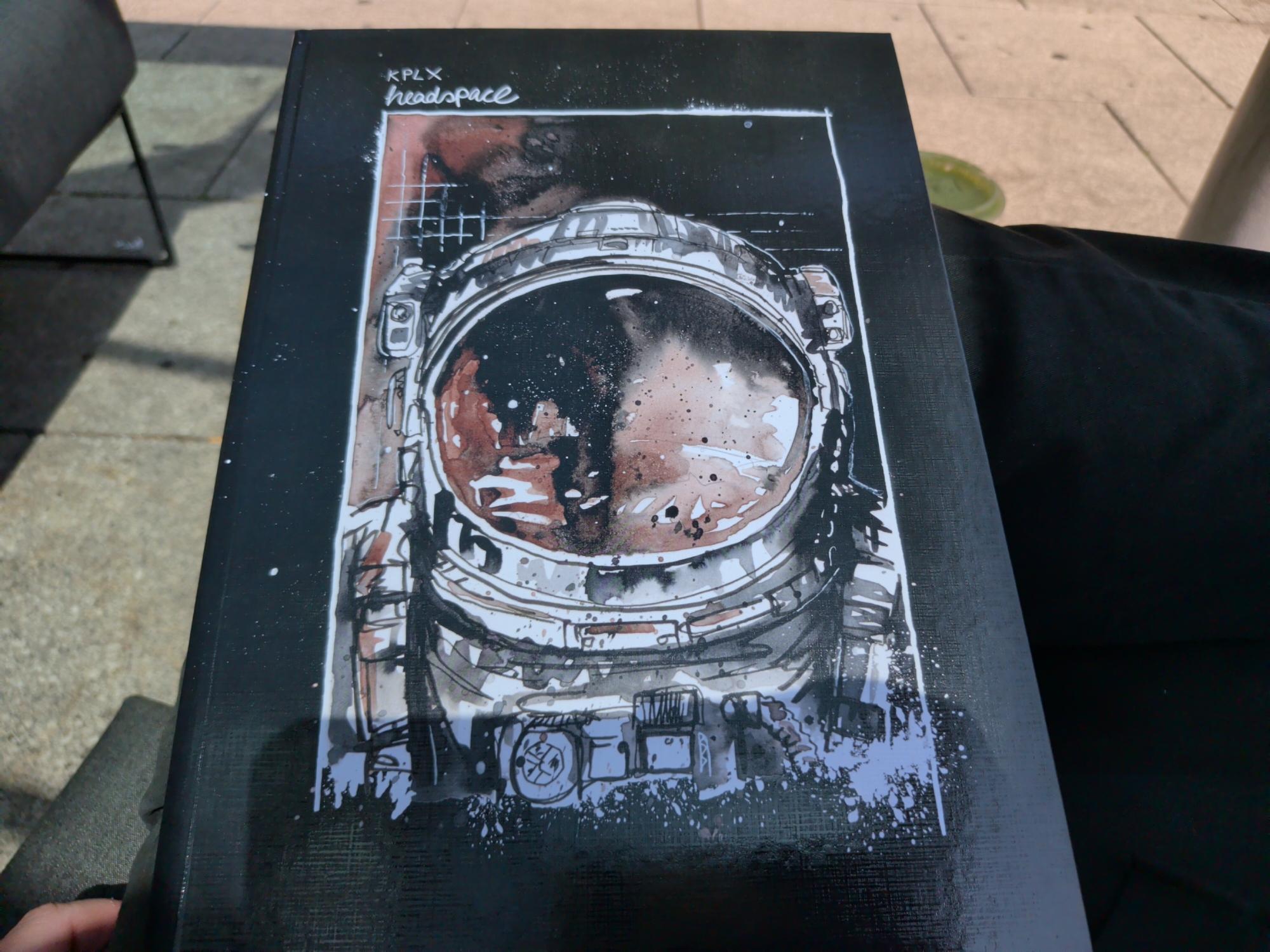 Ein schwarzes Buch mit einem Astronautenhelm auf dem Cover liegt auf meinen verschränkten Beinen.beim Hintergrund ein grünliches Getränk und ein wenig Sonnenschein.