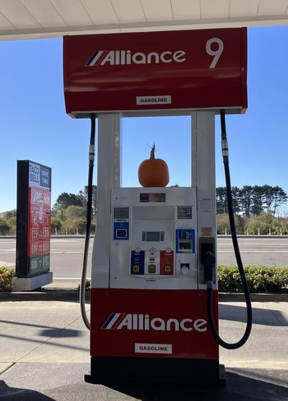 A gas station pump with a small pumpkin on top, on a day with a clear blue sky. In the background you can see the road the gas station is on and the sign listing the prices — although you can barely read the prices, given the angle and the distance. It's obvious the prices per gallon of all three grades of gasoline start with a "6" though, with $6.29 and $6.49 being two of them. Those are the cash prices; the credit prices are in a second column which you can't read.