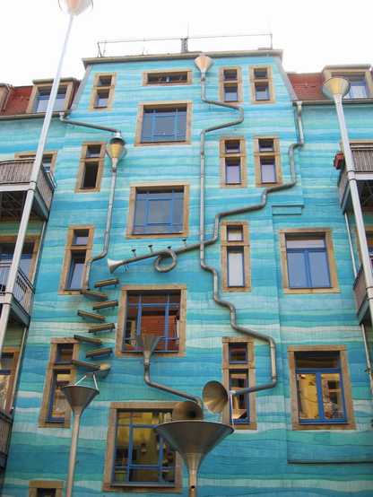 Photo of a facade of a multi-storey residential building in the Kunsthofpassage in Dresden. The Kunsthofpassage is a complex of buildings  with courtyards containing interesting artwork and decorations. This facade is painted in various shades of blue and the roof drainage pipes come down the facade bent and twisted, in part mimicking brass instruments, so when it rains they produce  interesting sounds.