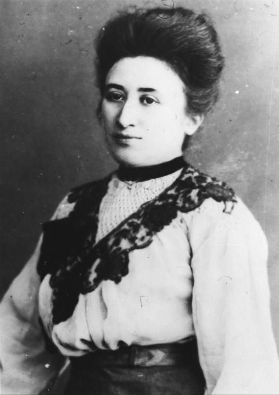 Image en noir et blanc de Rosa Luxemburg. Elle porte une blouse blanche ample avec des garnitures noires décoratives et une ceinture sombre est visible autour de sa taille. L’arrière-plan de l’image est sombre et uni.