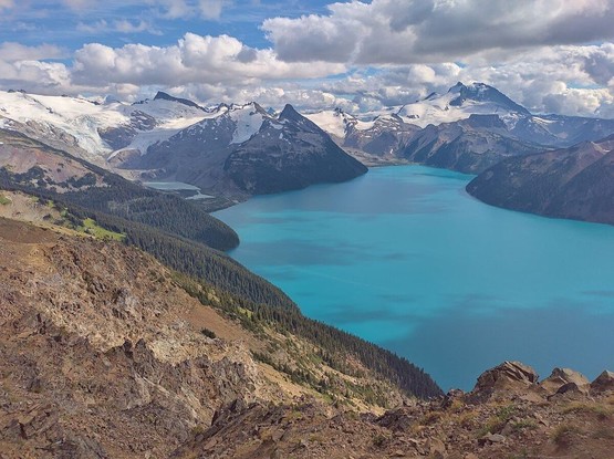 Vue aérienne du lac Garibaldi en Colombie-Britannique, mettant en valeur ses eaux turquoise entourées de montagnes majestueuses aux sommets enneigés, sous un ciel nuageux.