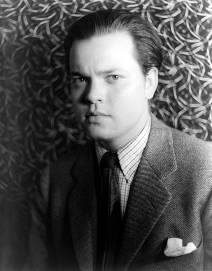 Photo d'Orson Welles en noir et blanc, portant un costume et une cravate. Il porte une chemise à carreaux et une veste sombre avec une pochette