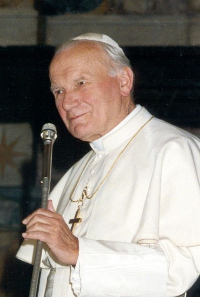 Photographie de Jean-Paul II. Il porte une robe blanche religieuse et un chapeau blanc. Il tient un microphone dans sa main. Un collier avec une croix dorée est visible autour de son cou, pendu sur la robe blanche. L’arrière-plan est sombre, ce qui met en évidence la personne en blanc.