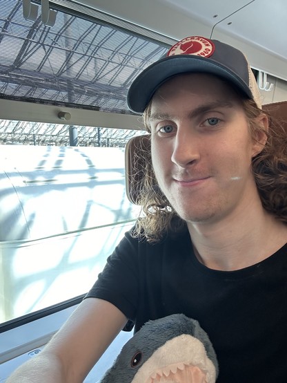 selfie of me with a black t-shirt and a hat, sitting next to a window on a VR ekstra (~first class) seat on an intercity train. i'm smiling, holding a blåhaj and in the window you can just see the roof of a VR Eil/Eilf loco-hauled commuter rail car getting ready for its singular run of the day (that I still need to ride at some point before they're retired because why not)