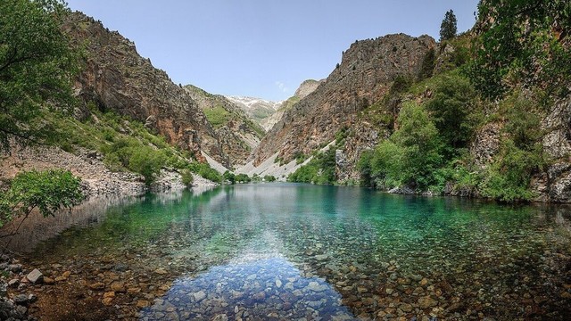 Photo d’un lac aux eaux turquoise cristallines dans la province de Tachkent en Ouzbékistan, niché dans une vallée entourée de falaises escarpées et de montagnes verdoyantes. La surface du lac reflète le paysage environnant, et le fond rocheux est visible à travers l’eau transparente. La végétation luxuriante orne les pentes des montagnes, témoignant d’un riche habitat naturel. 