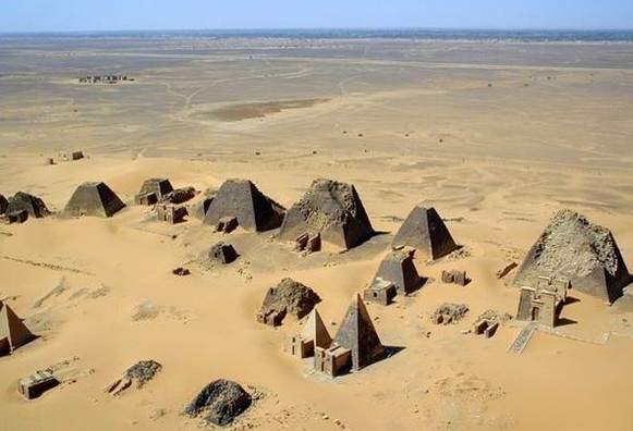 Vue aérienne du site archéologique de Méroé, montrant un groupe de pyramides nubiennes aux sommets tronqués et bases carrées, éparpillées sur une étendue de sable doré. Le ciel est clair et la perspective offre une vue sur l’horizon désertique lointain.