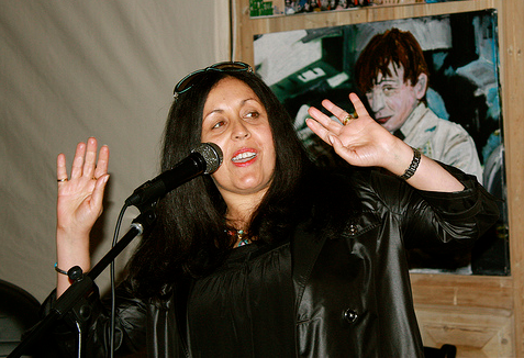 Photo de Poly Styrene, en train de parler dans un microphone, les mains levées. Elle porte une veste en cuir noire. En arrière-plan, il y a une image accrochée au mur. Le cadre semble être à l’intérieur avec des murs en bois.