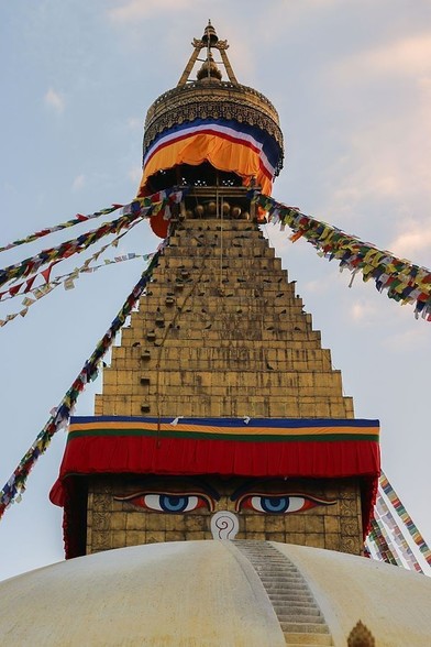 Photo du sommet du stupa de Bodnath au Népal. La flèche dorée emblématique du stupa se dresse contre un ciel bleu dégagé. Des drapeaux de prière colorés s’étendent depuis la flèche, créant un affichage vibrant. En dessous de la flèche, le harmika carré du stupa est visible, orné des yeux peints de Bouddha regardant vers l’extérieur, symbolisant l’omniscience. Le harmika est drapé d’un tissu orange, ajoutant à la sacralité et à l’aspect cérémoniel de la structure. La base en forme de dôme du stupa apparaît en bas de l’image, partiellement masquée mais toujours reconnaissable par sa forme arrondie.