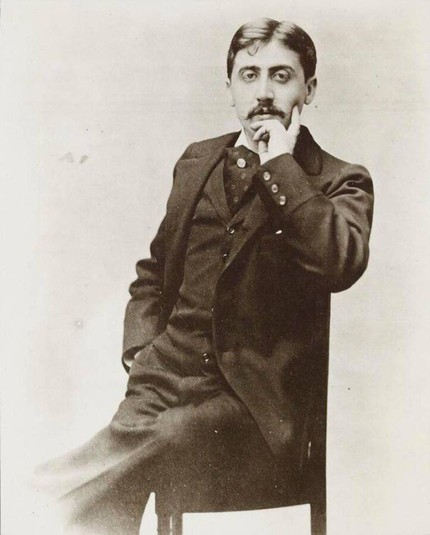 Photographie en noir et blanc de Marcel Proust. Il est assis sur une chaise, vêtu d’un costume sombre avec un gilet et une cravate. Sa posture est droite et formelle. L’image est claire et met l’accent sur Marcel Proust au premier plan.