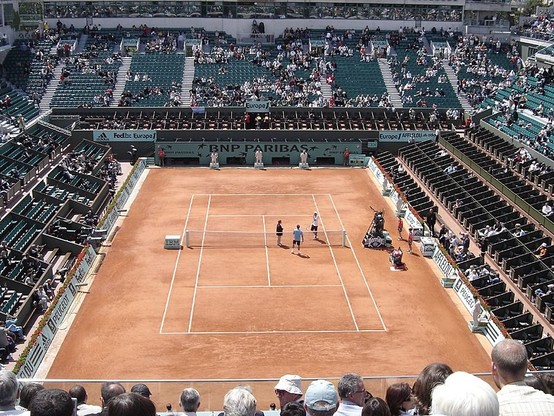 Photo du Court Philippe-Chatrier au stade Roland-Garros à Paris, mettant en avant un court de tennis en terre battue avec des lignes de délimitation blanches et un filet au milieu. Les gradins sont remplis de spectateurs, et plusieurs personnes sont présents sur le court.