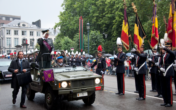 Photo montrant l'ancien roi des Belges Albert II, en uniforme, debout dans un véhicule militaire vert, saluant la foule ainsi que des doldats lors d’un défilé. Des officiers en uniforme portant des drapeaux sont alignés, et une foule de spectateurs observe en arrière-plan. Des bâtiments et des arbres bordent la rue.