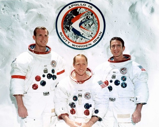 Photographie de l’équipe d’Apollo 15, composée de trois astronautes en combinaisons spatiales blanches, debout côte à côte devant un fond représentant la surface lunaire. Un logo circulaire d’Apollo 15 est visible au-dessus d’eux, affichant ‘APOLLO 15 SCOTT WORDEN IRWIN’.