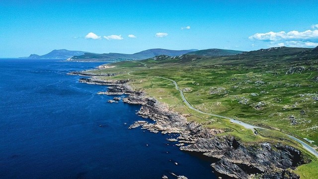 Highly commended photograph from WLE in Ireland 2023 by User:Kiran Madhusudhanan of the Achill Islands in County Mayo. An aerial shot of deep blue waters off a grassy coast, a winding road curves along the shore and a bright blue sky is overhead.