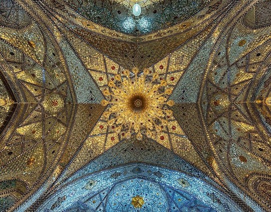 Photo du plafond du Chabestan du sanctuaire de Fatima Masoumeh à Qom, en Iran, avec un design islamique complexe et floral. Dominé par des tons de bleu avec des accents dorés et blancs, le plafond central comporte un grand lustre orné, entouré d’un motif de rayons de soleil. Ce motif se déploie ensuite en sections plus petites, richement décorées de mosaïques, de miroirs et de calligraphie, créant un motif symétrique visuellement saisissant. Le regard est attiré vers le haut, vers cette œuvre d’art magnifique. 
