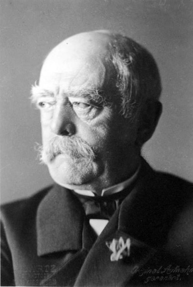 Photographie en noir et blanc de Bismarck, le crâne dégarni, vêtu d’un costume sombre avec un nœud papillon et une broche décorative sur le revers.