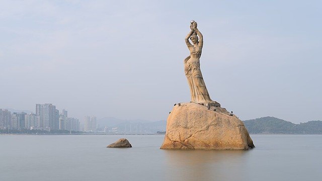 Photo de La Fille du Pêcheur est une statue en bronze érigée en 1982, devenue l’emblème de la ville chinoise de Zhuhai. La statue représente une femme debout sur un rocher au milieu d’eaux calmes. Elle tient gracieusement une perle au-dessus de sa tête, symbolisant l’histoire de Zhuhai liée à la pêche aux perles. En arrière-plan, un horizon flou de bâtiments modernes contraste avec la silhouette intemporelle de la statue. Le ciel est couvert, créant une atmosphère sereine.