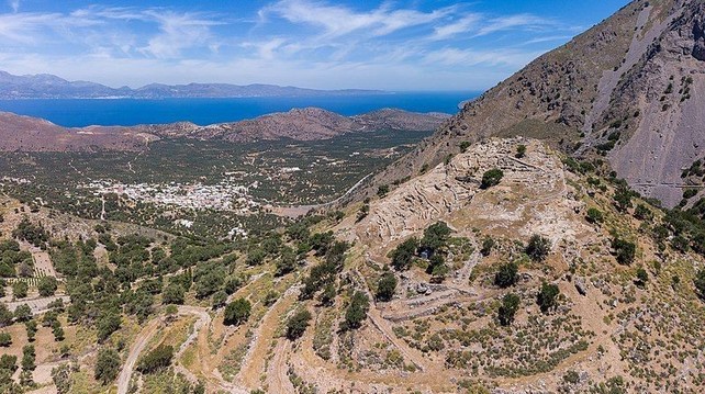 Vue aérienne du village de Kavoúsi et du golfe de Mirabello en Crète, avec le site archéologique d’Azoria sur la colline au premier plan. L’image montre un contraste entre la verdure luxuriante entourant le village et la colline aride et rocheuse. Des chemins de terre sinueux traversent le paysage, menant aux vestiges des structures anciennes sur les pentes d’Azoria. En arrière-plan, les eaux bleues et sereines du golfe de Mirabello ajoutent à la beauté de l’ensemble sous un ciel dégagé.