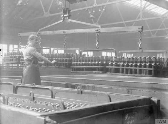 Photographie en noir et blanc de 1918 montrant le processus de pasteurisation dans une brasserie du comté de Cheshire. Une ouvrière se tient devant de nombreuses bouteilles sur des tapis roulants, avec des équipements industriels visibles en arrière-plan. L’usine spacieuse est éclairée par la lumière naturelle provenant de grandes fenêtres.