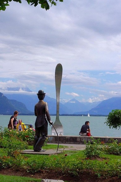 Statue de Charlot, de dos, face à la Fourchette de Vevey, avec le lac Léman et les montagnes en arrière-plan. Le ciel est partiellement nuageux et des promeneurs profitent du bord du lac.